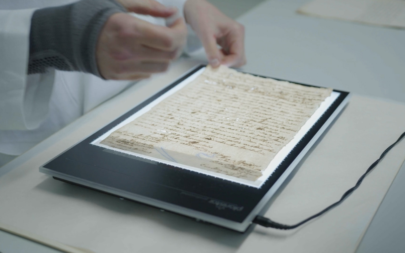 Filmstill aus »Digitizing Droste«. Der Film zeigt den Prozess des Digitalisierens von Handschriften im LWL-Archivamt. Das Filmstill zeigt einen Scanner, mit erleuchteter Oberfläche und einer Handschrift Drostes darauf. Eine mitarbeitende Person des Archivamts hält mit der linken Hand das Blatt an einer Ecke fest.