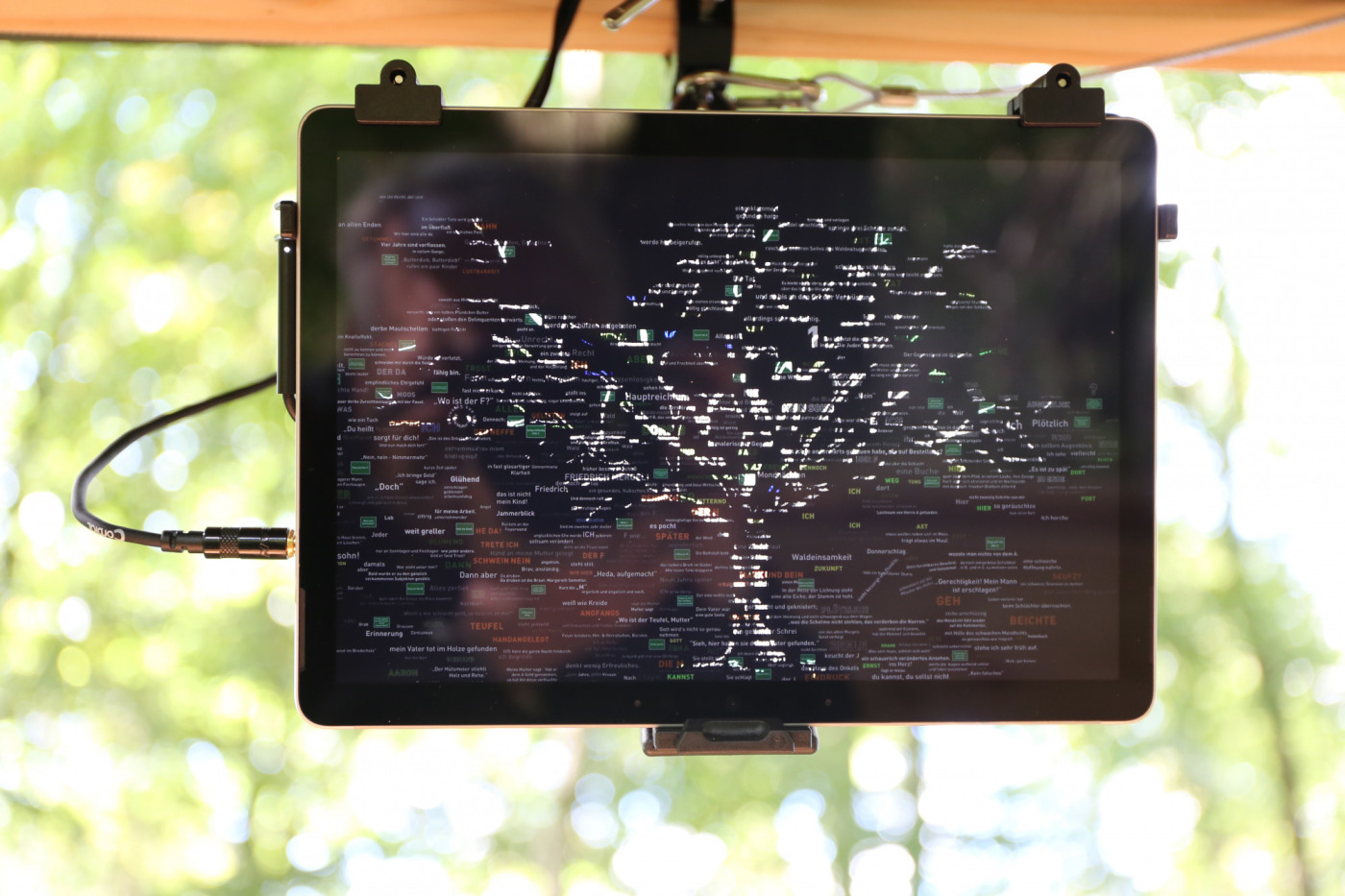 An der Decke eines Hochsitz befestigtes Tablet. Besucher*innen, die sich auf den Hochsitz setzen, können es betrachten. Der Bildraum ist voll von waagerecht erscheinenden Begriffen in verschiedenen Größen. Die Begriffe sind unterschiedlich eingefärbt, sodass sich aus ihnen teilweise das Bild eines Baums ergibt.