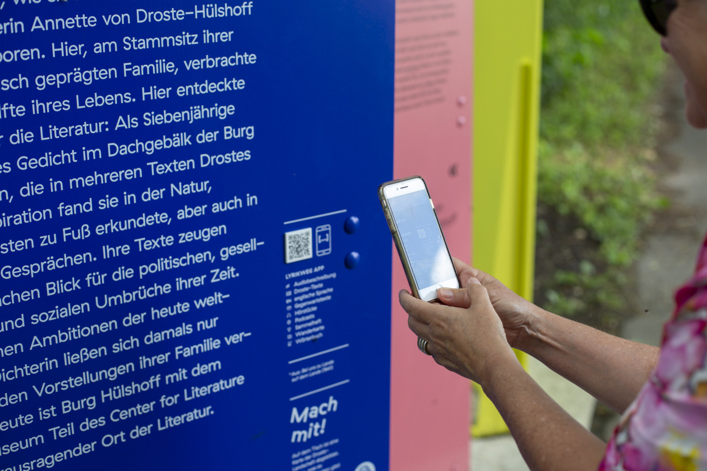 Foto, das zeigt, wie eine Person, die an einer Lyrikweg-Tour teilnimmt einen QR-Code mit dem Smartphone scannt. Der QR-Code befindet sich auf einer Tafel an einer der Lyrikweg-Stationen. An jeder Station befinden sich Tafeln mit Informationen zu Annette von Droste-H&uuml;lshoff, ihrem Werk und Leben.
