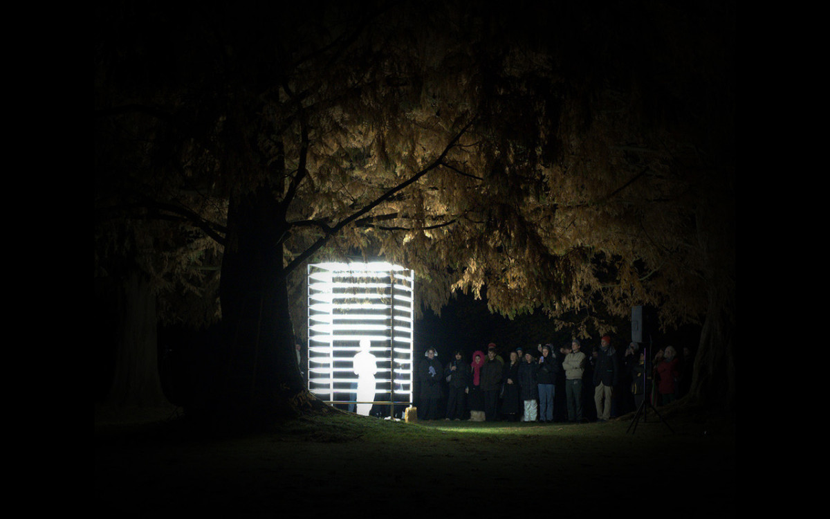 In the dark park, a cube made of neon tubes can be seen in which an illuminated person is standing. Several spectators are standing in front of it on the right.