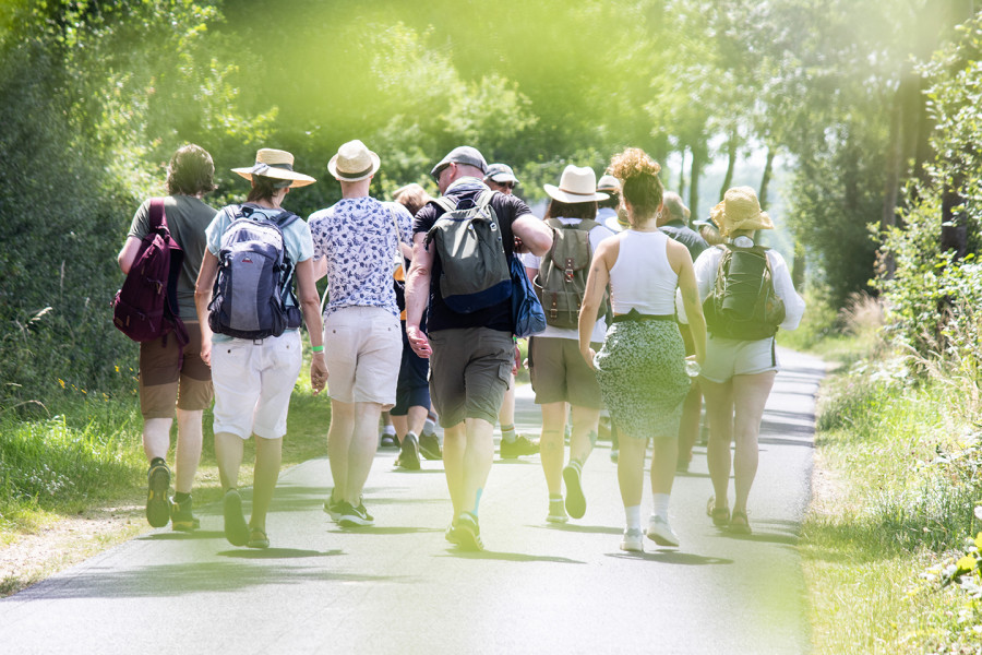 A group of people can be seen from behind walking together along a path. It is summer weather, people are wearing shorts and sun hats.