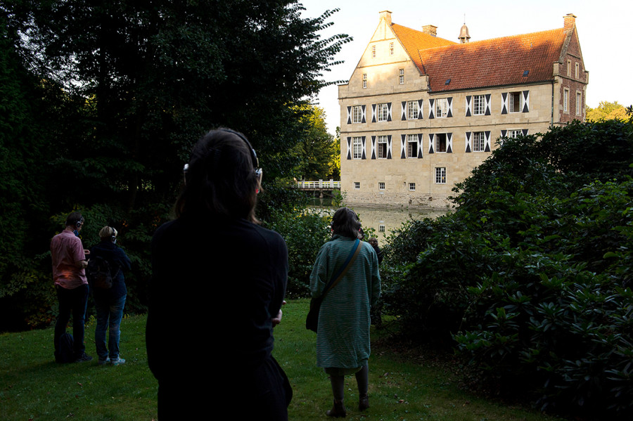 Four people with headphones are standing with their backs to the viewer on a field, which is bordered by bushes and a moat in front. In the background, the manor house of Burg Hülshoff can be seen in the evening sun.