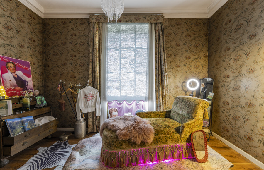 A room lined with antique fabric wallpaper, in which a chaise longue with sheepskin draped on it stands in the middle. Behind it is a window with a black blind on which white handwriting can be seen. Around the chaise longue are various objects, from a mirror, selfie lamp, fan jumpers, ski poles, a display case with a record on top, on the floor a zebra skin and a kind of carpet made from a map.
