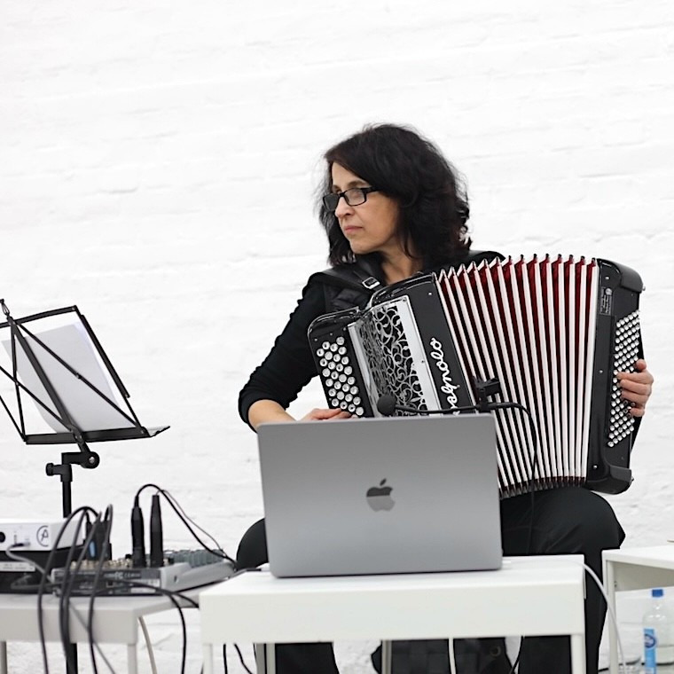 Klang- und Medienkünstlerin Anna Kreysing sitzt an einem weißen Tisch und spielt Akkordeon. Vor ihr stehen ein Laptop, ein Notenständer und Audiotechnik. Der Hintergrund ist eine weiße Backsteinwand. Foto: © Hannah Neander