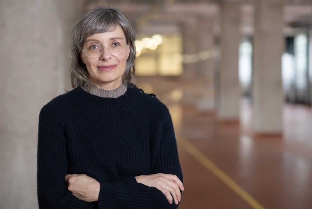 Das Bild zeigt Katrin Mayer mit verschränkten Armen in einer Halle mit Säulen