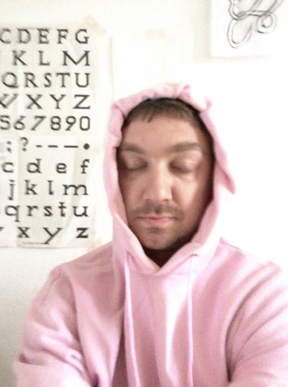 Max Grau hat die Augen geschlossen und trägt einen rosa Pullover mit Kapuze. Hinter ihm hängt ein Plakat mit Buchstaben.