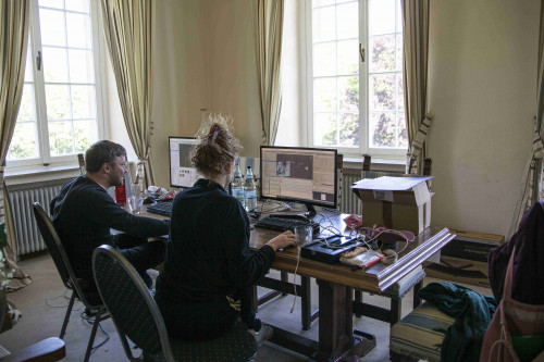 Fabian Raith und Natalie Sontopski von den Code Girls sitzen in der Burg Hülshoff an einem großen Holztisch mit Bildschirmen, Tastauren und vielen Kabeln.mit