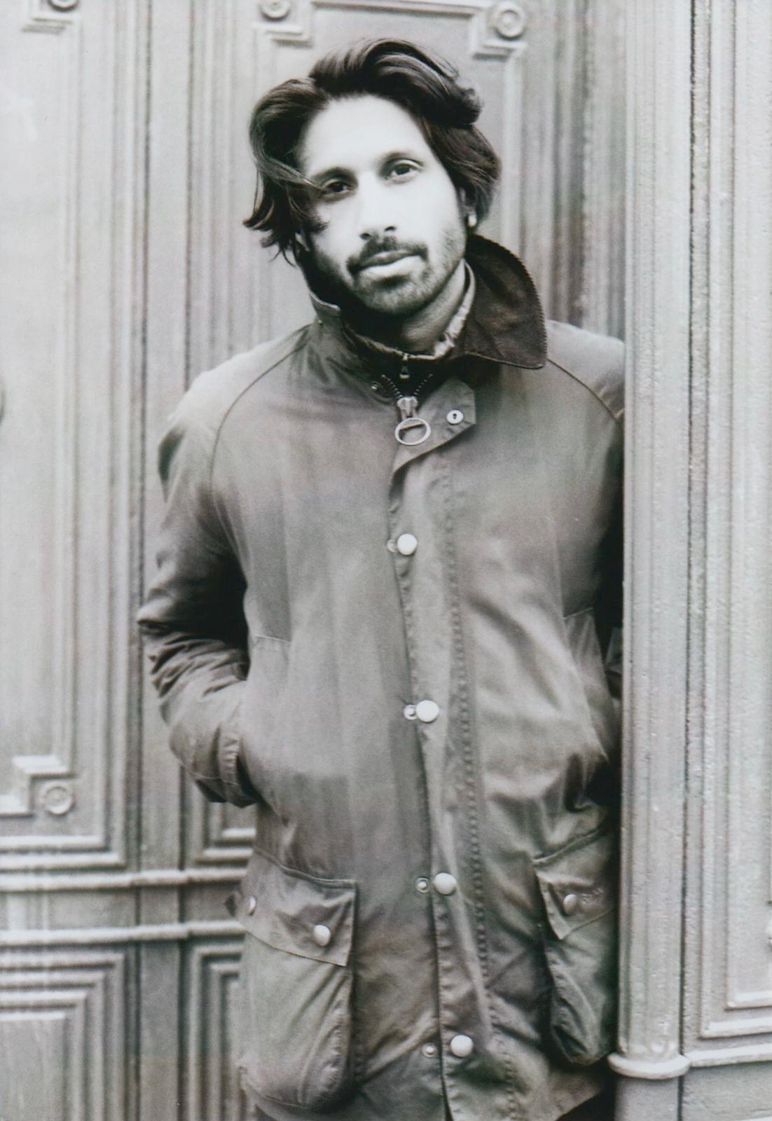 Ein schwarz-weiß Foto von Ralph Tharayil, der mit den Händen in der Jackentasche in einer Tür steht und in die Kamera blickt.