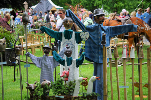 Handgefertigte Puppen stehen auf einer grünen Fläche, im Hintergrund sieht mensch die Besucher*innen der Gartenträume 2019.