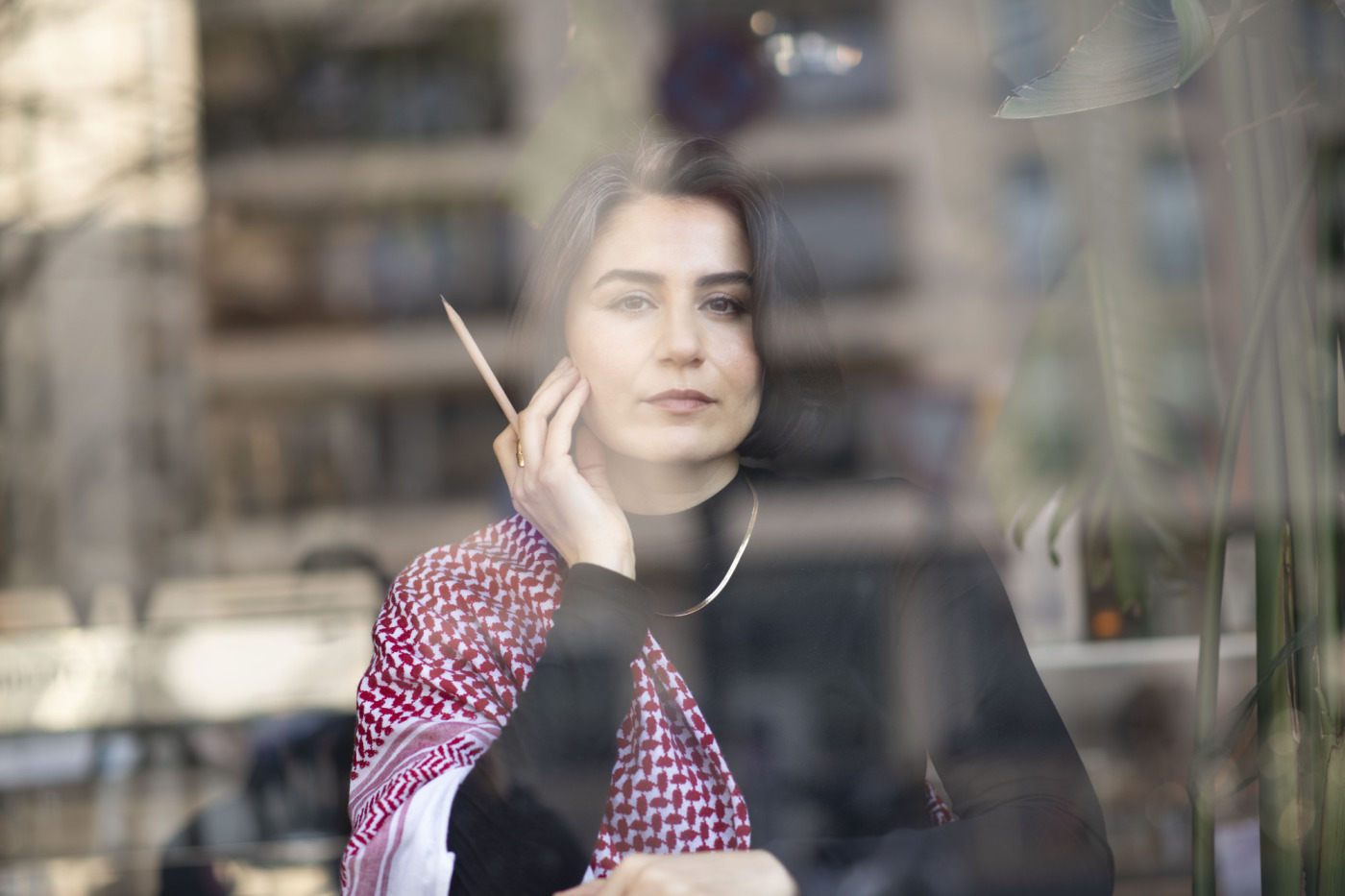 Ein Porträtfoto der Autorin Karosh Taha hinter einer Fensterscheibe zu sehen. Sie hält die Fingerspitzen ihrer rechten Hand an ihr Gesicht und einen Bleistift in derselben Hand. Ihre rechte Schulter ist mit einem Kufiya-Tuch bedeckt.