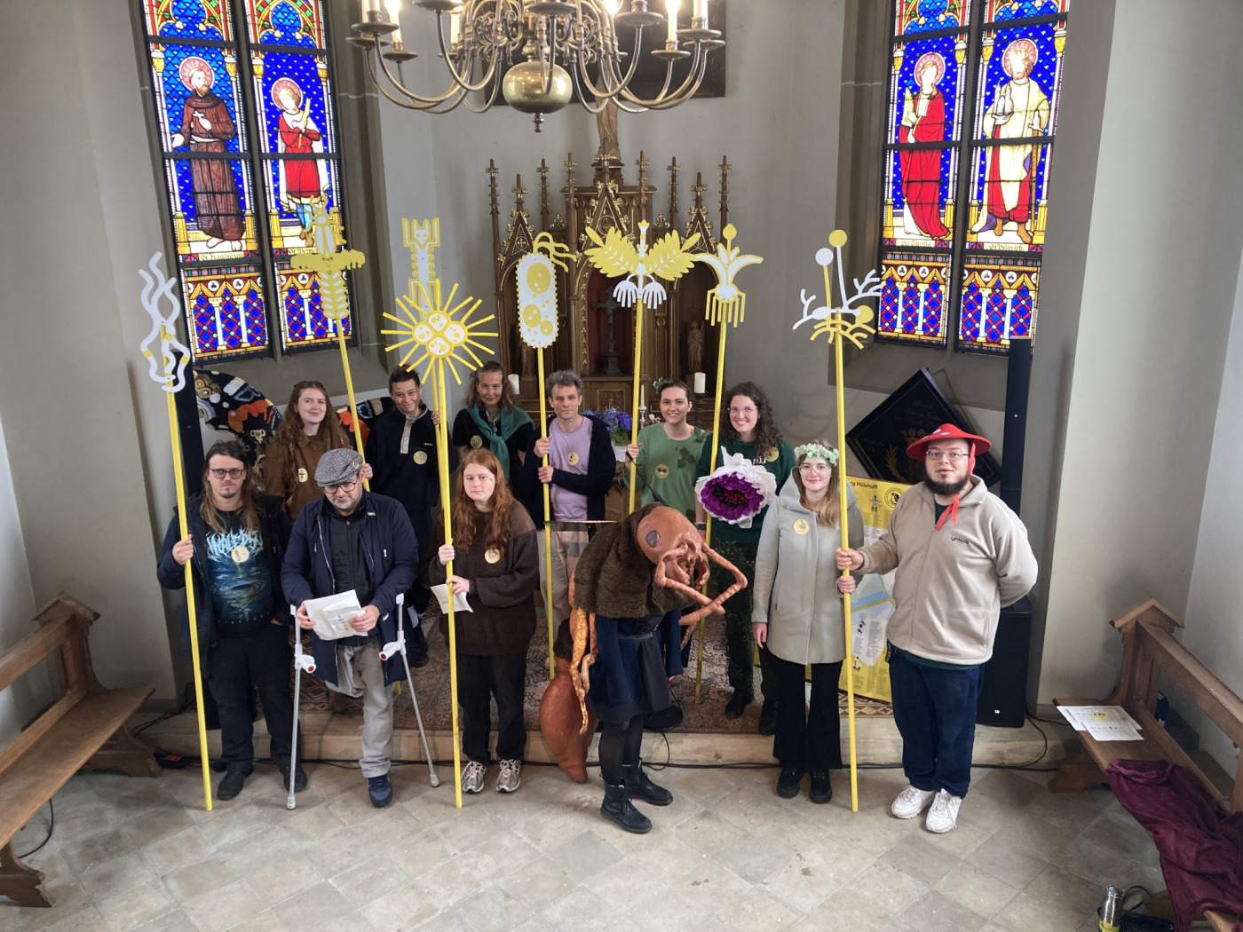 Foto des Kollektivs Club Real, das die Organismendemokratie darstellt. Die Mitglieder sind teilweise als unterschiedliche vertretene Organismen verkleidet und stehen mit symbolischen Stäben vor dem reich verzierten Altar der Kapelle von Burg Hülshoff.