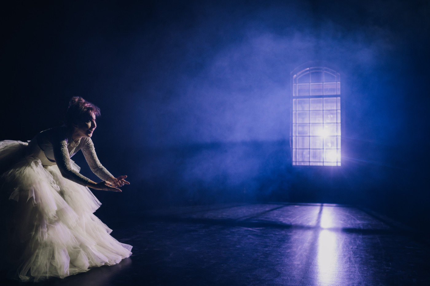 Regine Andratschke als Schauspielerin in der Rolle des Käthchen auf einer Bühne. Der Bühnenboden und die Umgebung ist tiefdunkel. Violettfarbenes Licht aus einem Fester flutet die Bühne in nebeliger mystischer Atmosphäre. Links im Bild zu sehen: Käthchen verbeugt sich gerade aus schauend und trägt ein Korsett mit bodenlangem Tüll Rock.
