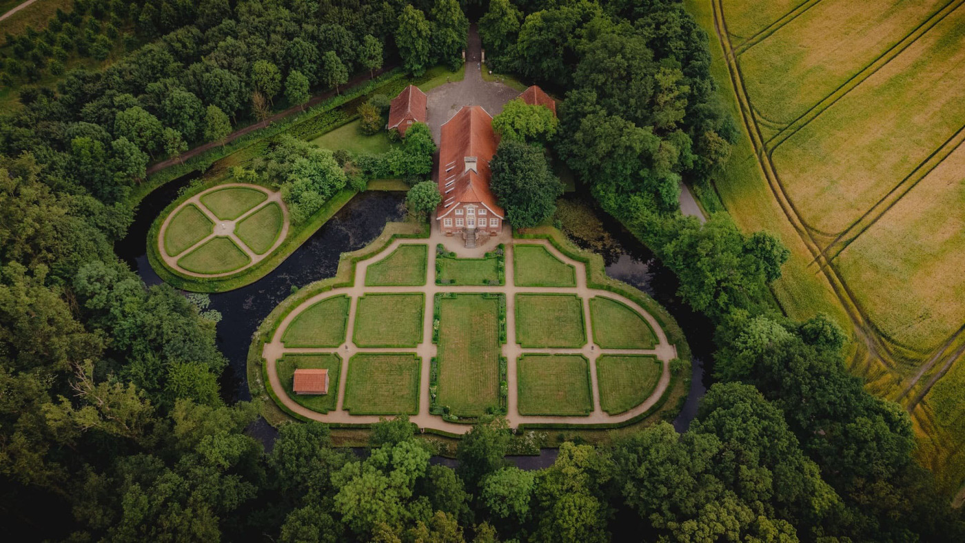 Haus Rüschhaus samt Gartenanlage und Umgebung aus der Vogelperspektive. Das Grundstück ist von Bäumen und angrenzend Feldern umgeben.