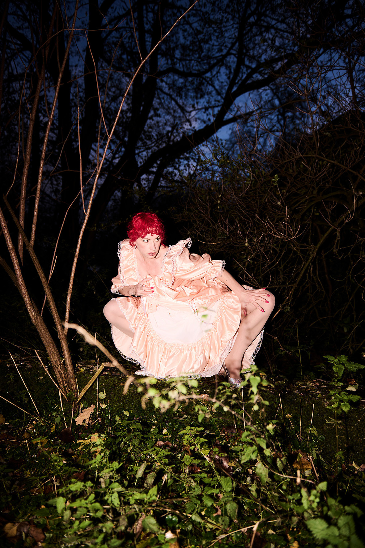 Szene bei Nacht zwischen Sträuchern und auf Waldboden. Eine Person mit roten Haaren, hellem, gerüschten Kleid und hohen Schuhen im Zentrum des Bilds. Die Person ist in die Knie gegangen und befindet sich in einer eher abwehrenden Haltung, mit skeptischem Blick aus dem Bildraum rechts heraus. Sie wird von der Kamera angeleuchtet.