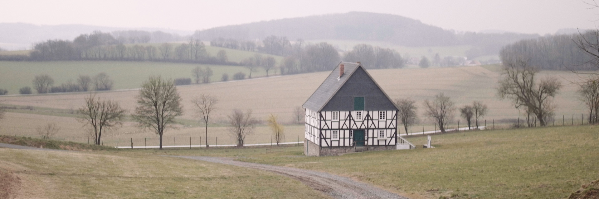 Foto, das ein Fachwerkhaus in einer h&uuml;geligen, weiten Landschaft zeigt. Das Haus ist alleinstehend, andere H&auml;user sind nicht zu sehen. Es f&uuml;hren Feldwege am Haus entlang.
Der Himmel ist zugezogen, die B&auml;ume sind nicht belaubt. Filmstill aus &raquo;The Geometry of Hope&laquo; von Daniel Laufer aus dem Jahr 2022.