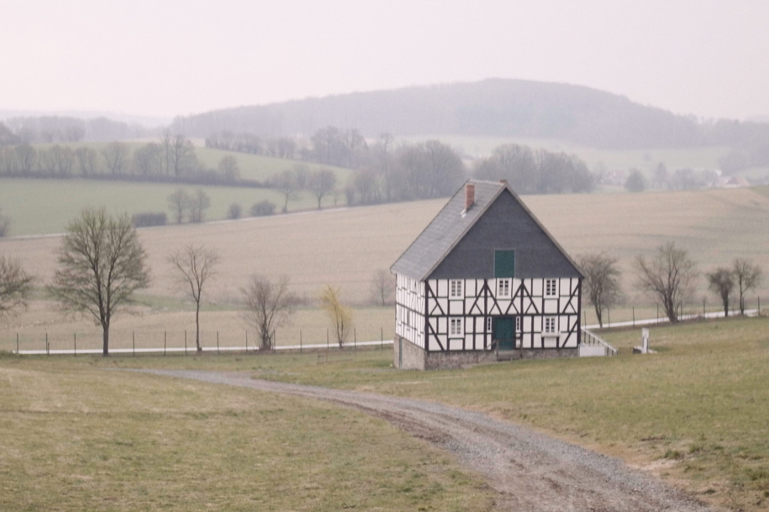 Foto, das ein Fachwerkhaus in einer h&uuml;geligen, weiten Landschaft zeigt. Das Haus ist alleinstehend, andere H&auml;user sind nicht zu sehen. Es f&uuml;hren Feldwege am Haus entlang.
Der Himmel ist zugezogen, die B&auml;ume sind nicht belaubt. Filmstill aus &raquo;The Geometry of Hope&laquo; von Daniel Laufer aus dem Jahr 2022.