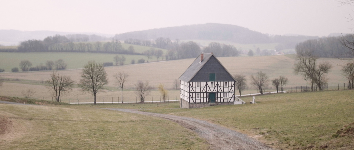 Foto, das ein Fachwerkhaus in einer h&uuml;geligen, weiten Landschaft zeigt. Das Haus ist alleinstehend, andere H&auml;user sind nicht zu sehen. Es f&uuml;hren Feldwege am Haus entlang.
Der Himmel ist zugezogen, die B&auml;ume sind nicht belaubt. Filmstill aus &raquo;The Geometry of Hope&laquo; von Daniel Laufer aus dem Jahr 2022.