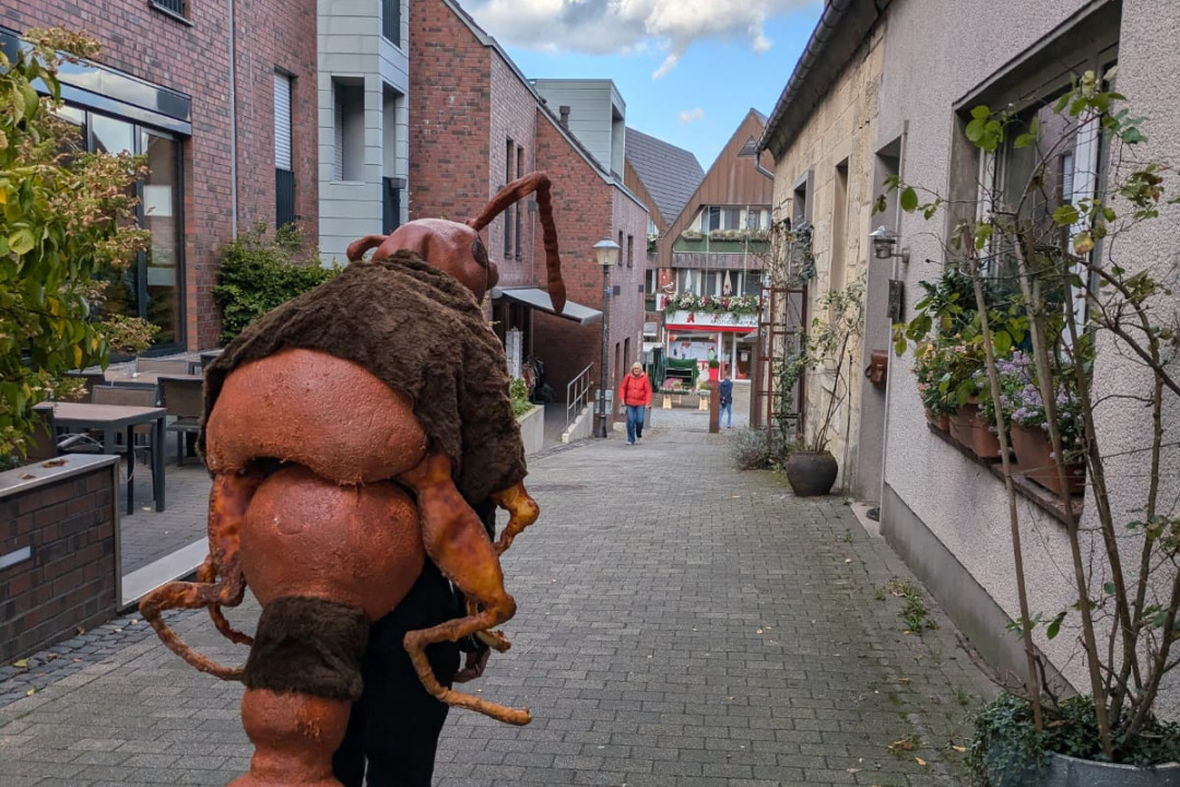Ein*e K&uuml;nstler*in der Gruppe Club Real in einer kleinen Gasse. Die Person wurde von hinten fotografiert. Sie tr&auml;gt das Kost&uuml;m einer Ameise. Der &raquo;K&ouml;rper&laquo; der Ameise ist wie ein Panzer von hinten auf die Person drauf gesetzt.