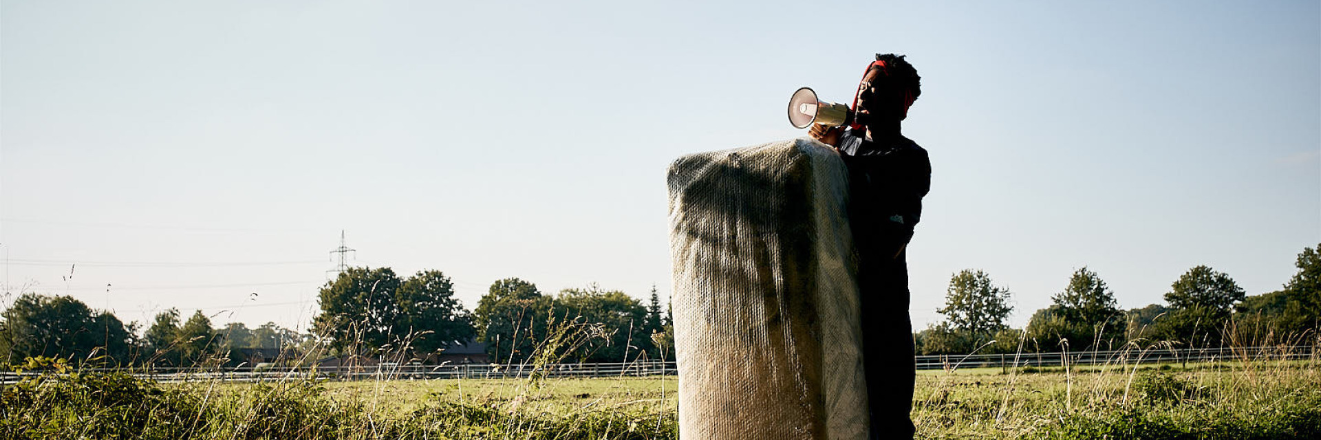 Foto von einem Halt der Radtour, die im Rahmen von »Westfailure - The Performance« statt gefunden hat. Das Foto zeigt eine Performance. Eine Person steht hinter einem großen, sack-ähnlichen Objekt und spricht etwas in ein Megaphon. Die Person steht auf einem Feldweg neben einer Wiese. Im Hintergrund befinden sich Bäume und Strommasten.