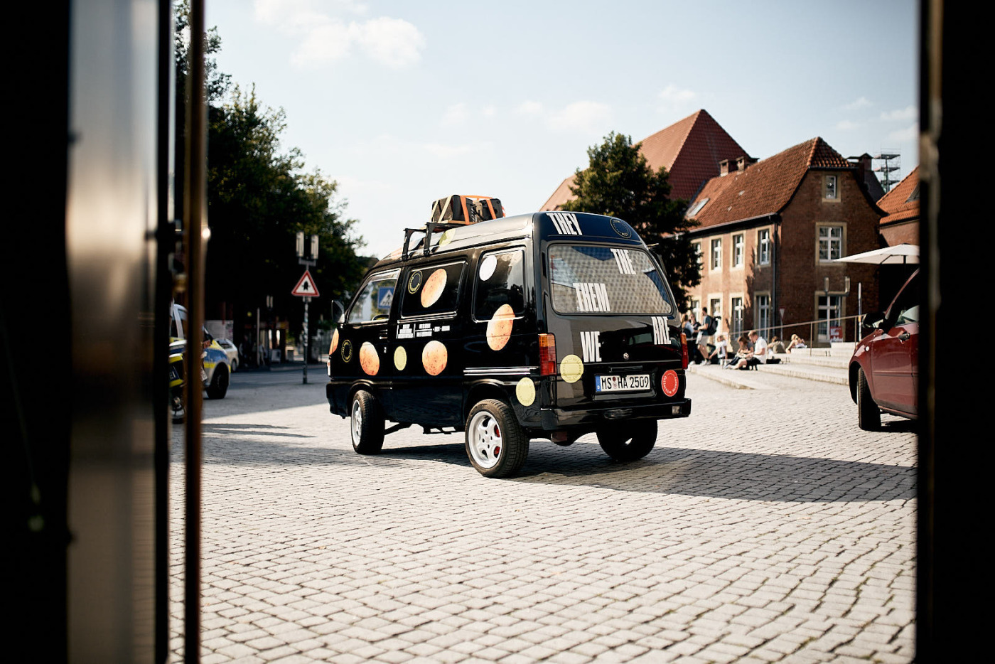 »Potato Propaganda Car« von Nástio Mosquito. Ein kleiner schwarzer Lieferwagen mit über das gesamte Auto verteilten Aufdrucken von Kartoffeln. Auf der Kofferraumklappe sind Begriffe wie »THEY« und »WE« aufgeklebt. Das Auto steht auf dem Platz vor dem Westfälischen Kunstverein in der Innenstadt von Münster.