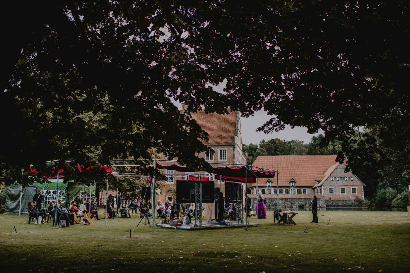 Outdoor-Bühnenaufbau mit Publikum im Park von Burg Hülshoff während des »Westopia« Festivals. Das Foto ist aus der Ferne aufgenommen. Im Hintergrund ist die Burg zu sehen.