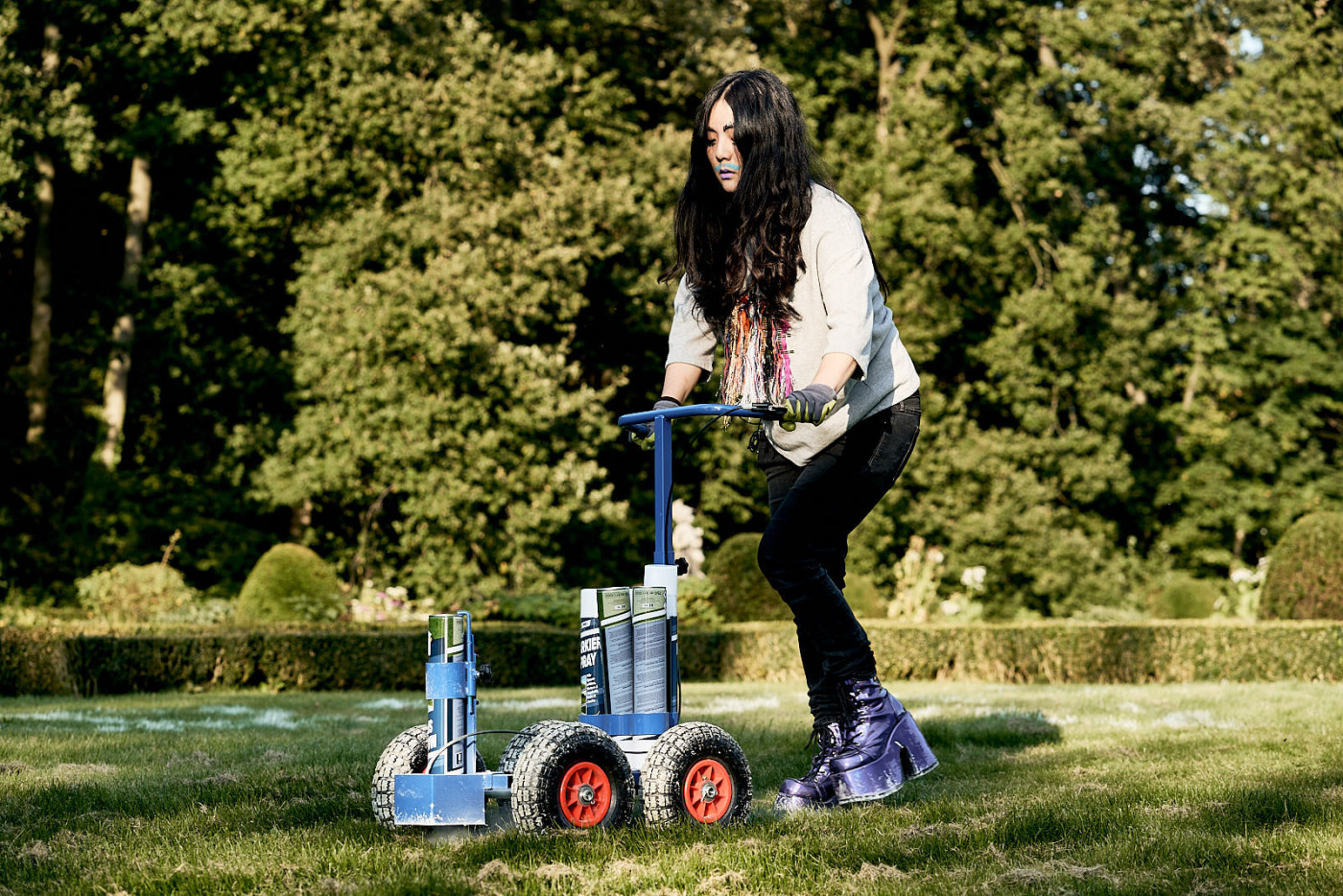 Die Choreographin Olivia Hyunsin Kim zeigt eine Performance im Garten von Haus Rüschhaus. Kim trägt neben Hose und Oberteil auch Arbeitshandschuhe und Plateauschuhe. Vor sich schiebt Kim ein Objekt, mit dem Markierungen auf der Grasfläche aufgebracht werden können.