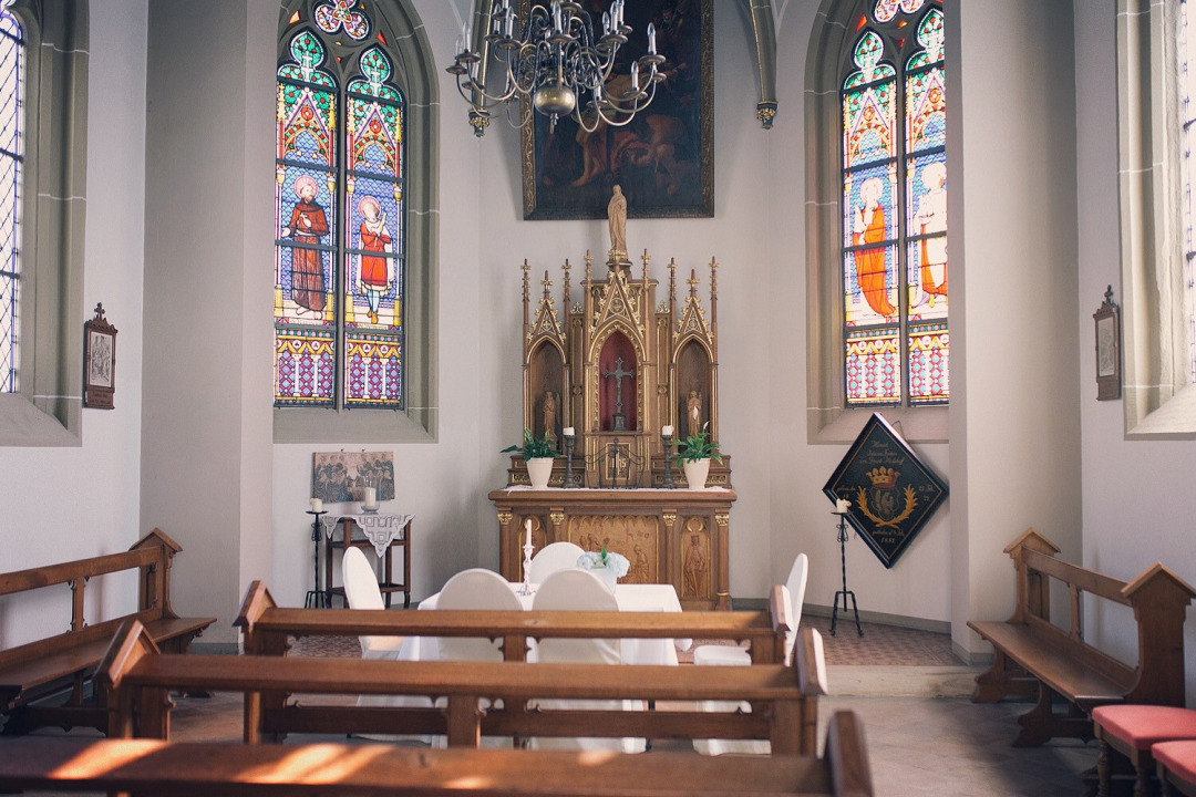 Innenraum der Kapelle auf Burg Hülshoff. Kirchenbänke, dahinter ein Tisch mit fünf Stühlen für die Trauung. Im Hintergrund ein hölzerner Altar. Mehrere bunte Kirchenfenster und ein Kronleuchter.