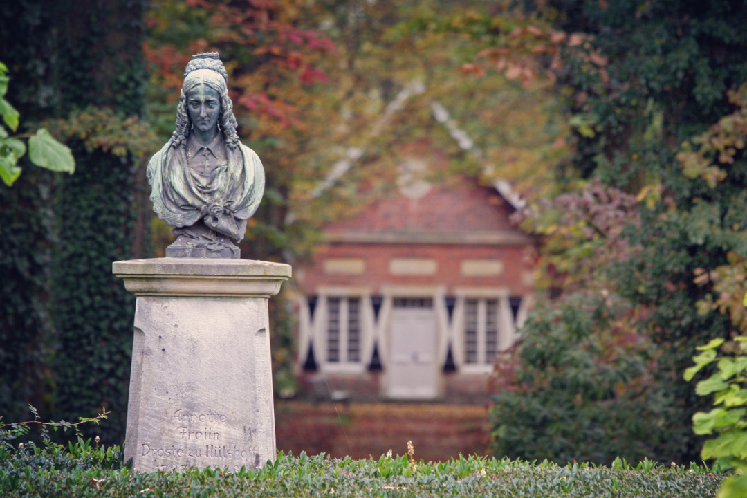 B&uuml;ste, die das Abbild von Annette von Droste-H&uuml;lshoff zeigt. Die B&uuml;ste steht auch einem Sockel im Park von Burg H&uuml;lshoff. Im Hintergrund liegt das &raquo;Teeh&auml;uschen&laquo;. Der Blick von Droste ist nach unten gerichtet. Ihre Haare sind, f&uuml;r die Zeit des Biedermeier typisch, am Hinterkopf hochgesteckt und an den Schl&auml;fen gelockt.