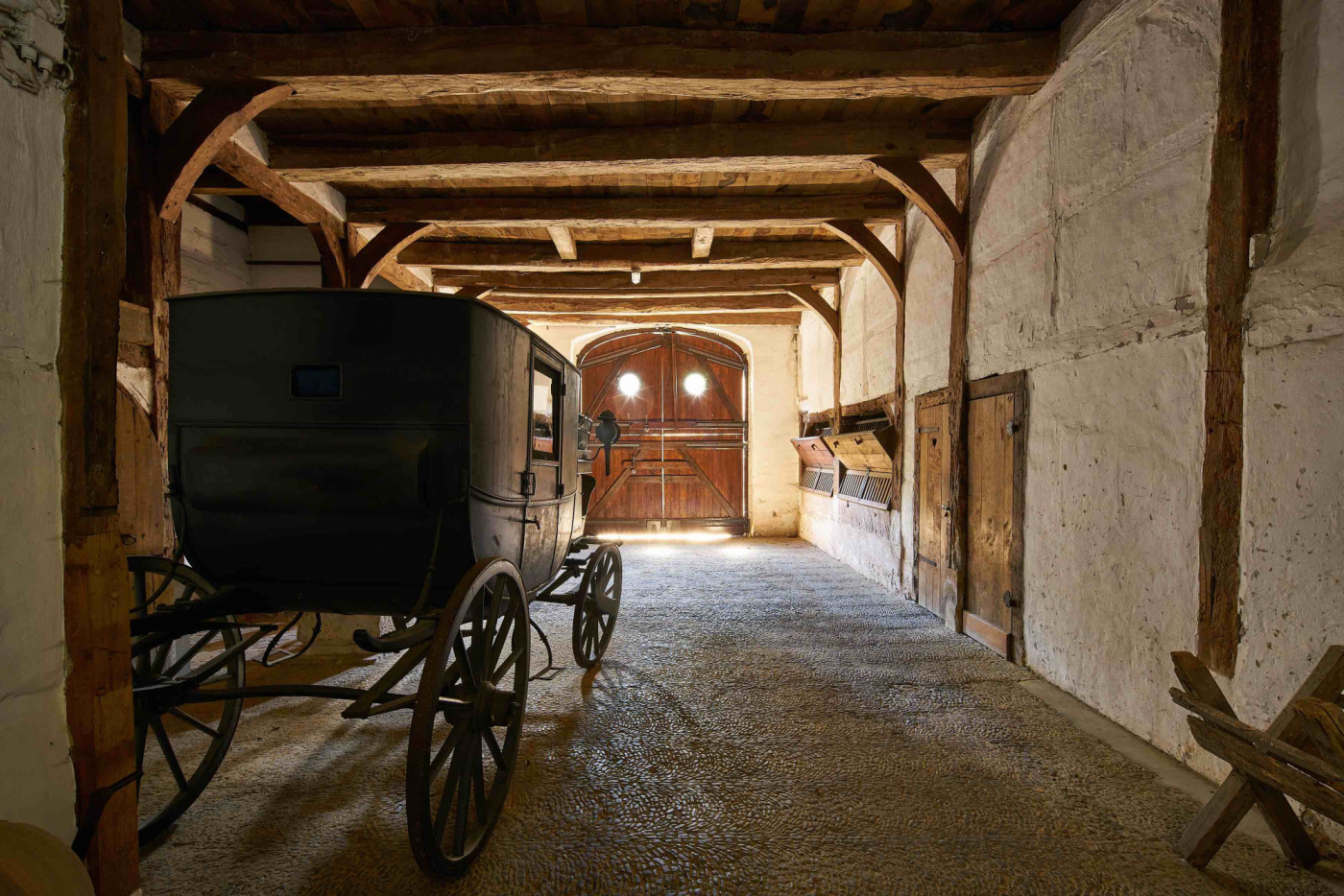 Kutsche, fotografiert von hinten in der &raquo;Alten Tenne&laquo; in Haus R&uuml;schhaus. Die Decke der Tenne wird von gro&szlig;en, dicken Holzbalken gest&uuml;tzt, die W&auml;nde sind wei&szlig; verputzt, an der rechten Seite des Raums sind alte Futtertr&ouml;ge zu sehen. Am Ende des Raums liegt ein gro&szlig;es Tor mit zwei runden Fenstern.