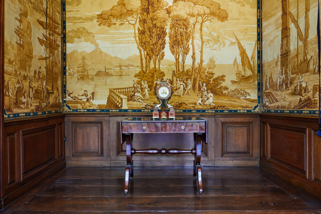 Foto, das im Inneren von Haus Rüschhaus, im »Italienischen Zimmer«, aufgenommen wurde. Die Wand ist mit einer Tapete verkleidet, die noch aus der Zeit der Familie von Droste-Hülshoff stammt und Szenen im Hafen von Neapel zeigt. Außerdem ein Tischchen mit einer alten Uhr.